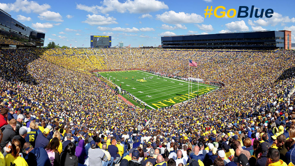 A shot of the University of Michigan Stadium with a crowd on football game day and the hastag Go Blue in the sky